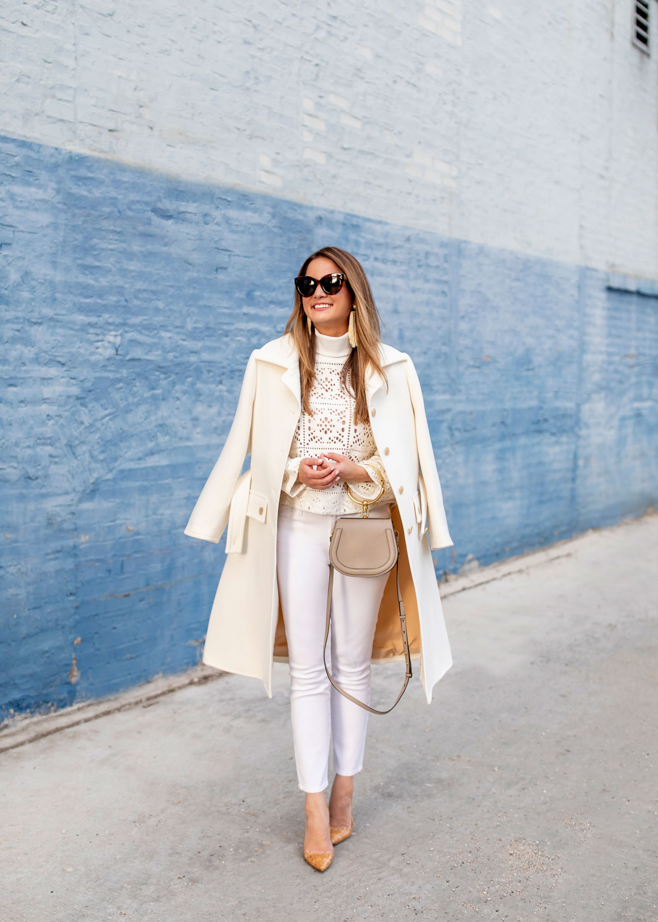 Woman in a cream coat over a patterned top with white skinny jeans and nude heels