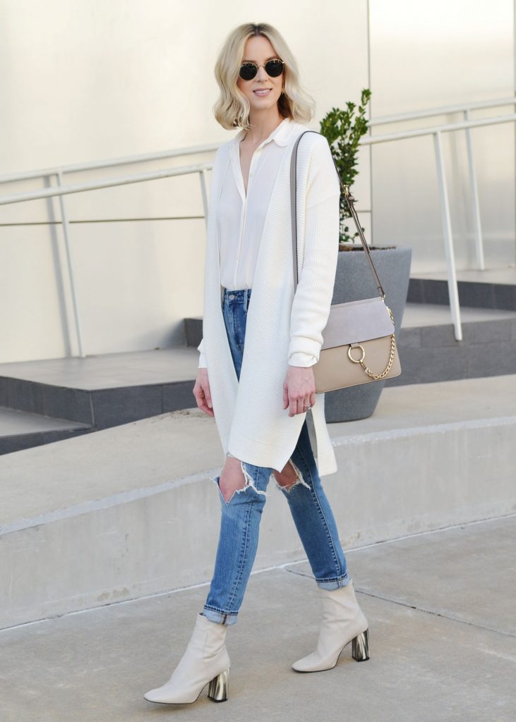Woman wearing a long cream cardigan over a white top with ripped jeans and white boots